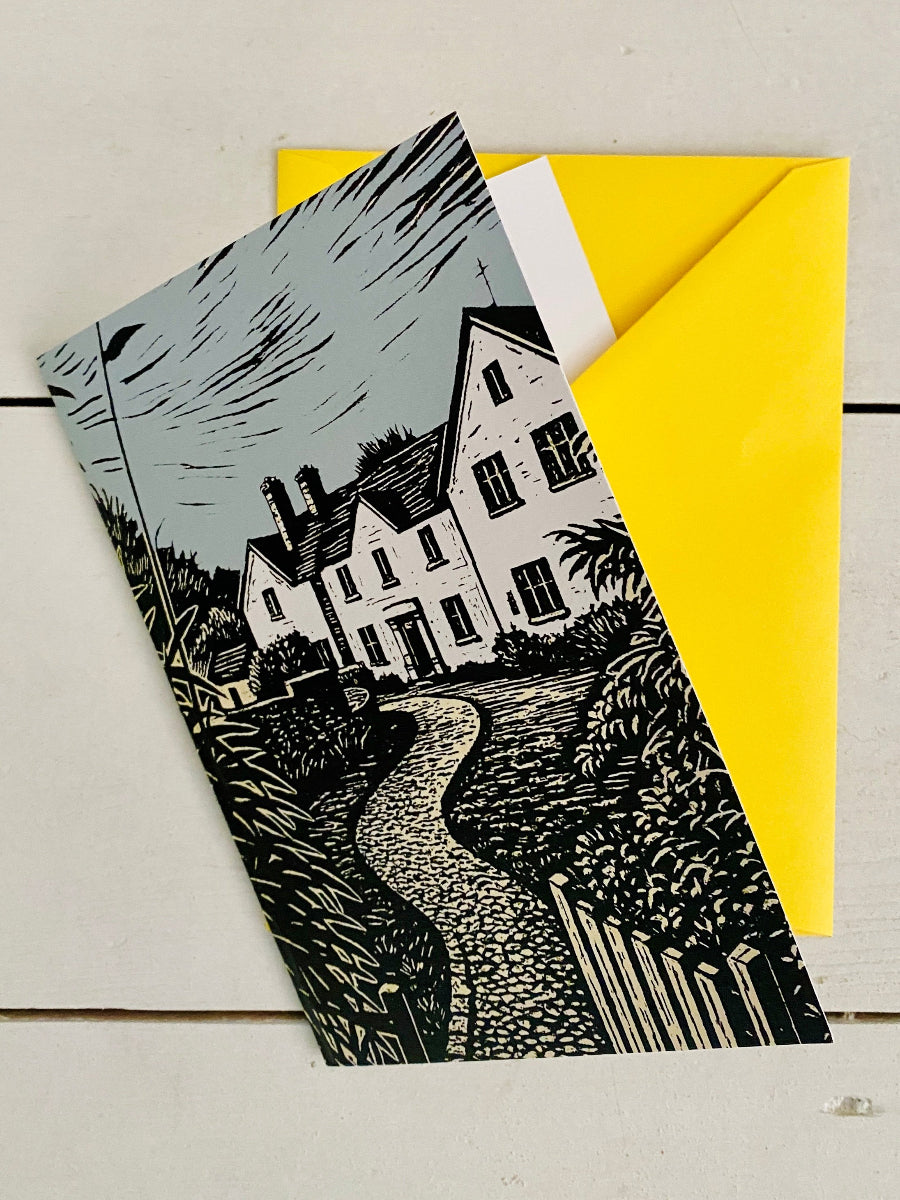 A printed card or art print featuring a black-and-white illustration of a house and winding path, placed over a bright yellow envelope on a light wood surface.