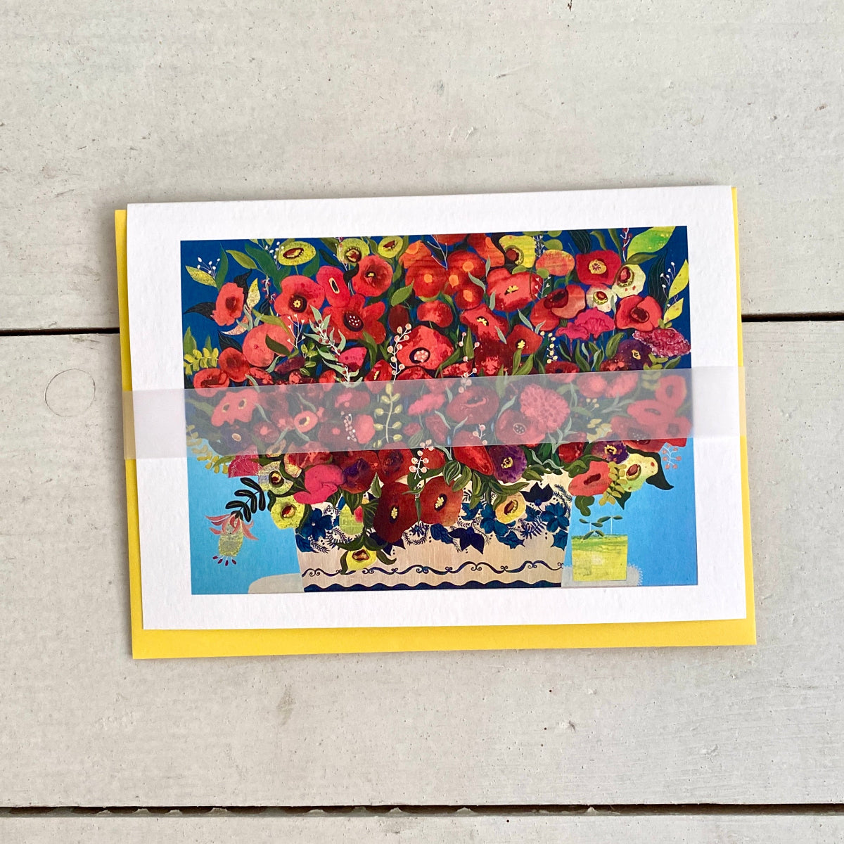 A rectangular greeting card featuring a colourful floral illustration with red poppies and other flowers in a blue vase, placed on a light gray wood surface with a yellow backing card visible around the edges.