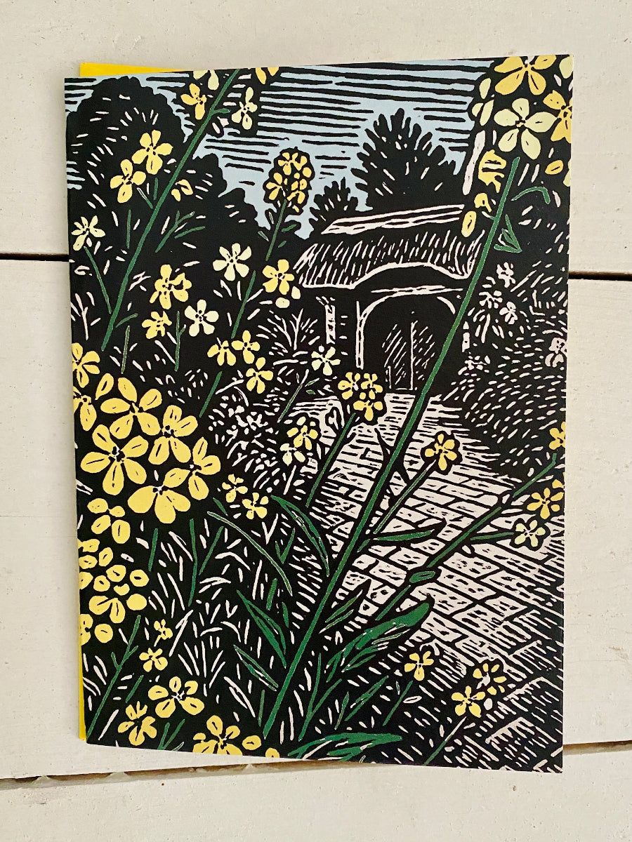An art card featuring a linocut-style illustration of yellow flowers and green stems in front of a stone path and small building, photographed against a light wood background.