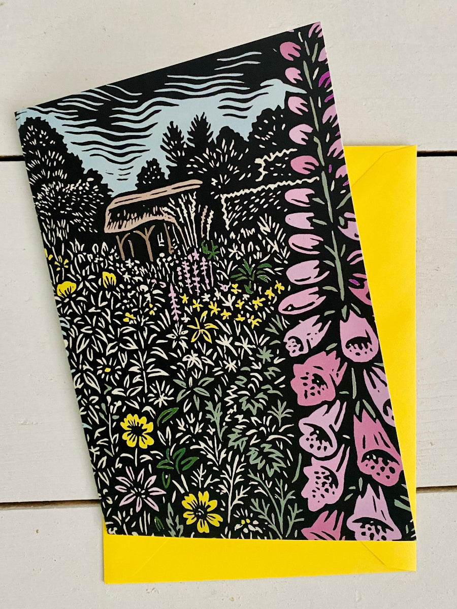 A square art print or card featuring a detailed floral and landscape illustration with a cottage, trees, and wildflowers, shown on a white wood background with a bright yellow envelope underneath.