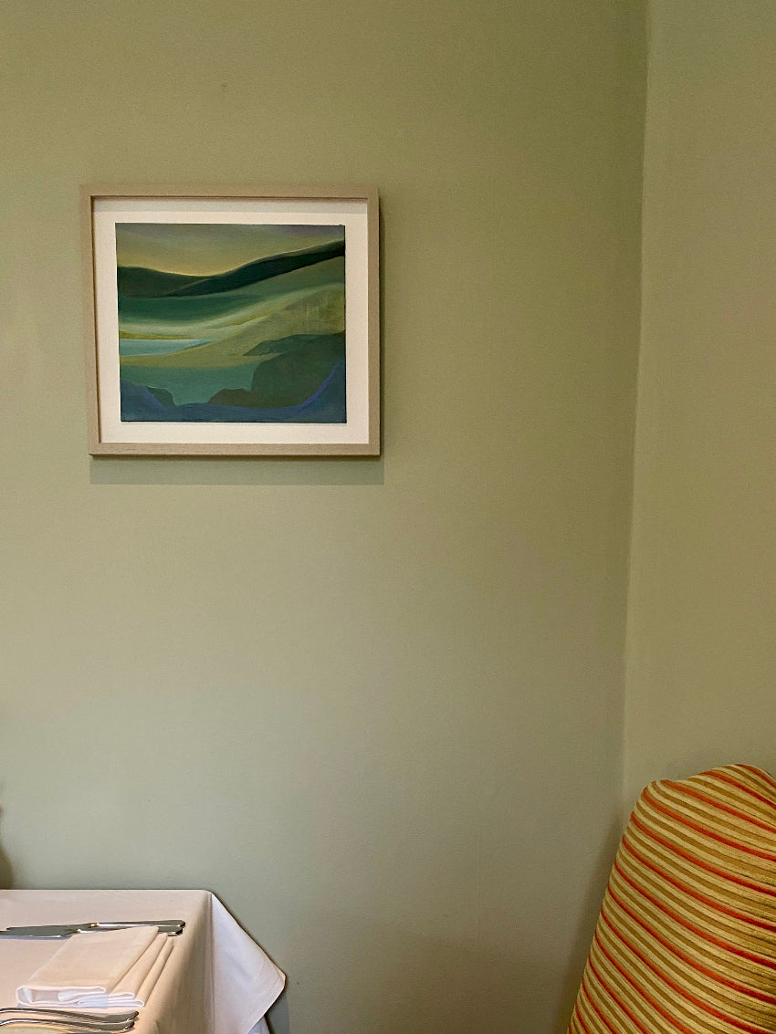 Framed abstract painting on a beige wall with a chair and table in the foreground.