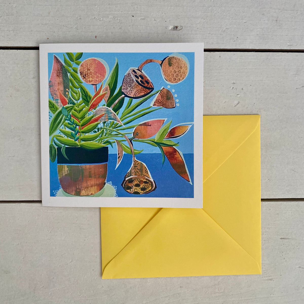 A square art card print featuring a colourful illustration of a potted plant with green leaves and brown seed pods against a bright blue background, placed on a light grey wood surface with a yellow envelope behind it.