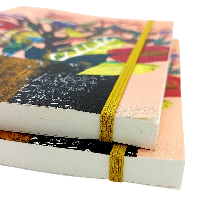 Two softcover sketchbooks with colourful abstract and botanical covers, stacked and secured with mustard yellow elastic bands, on a white background.