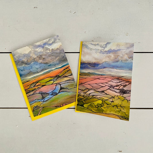 Two watercolor landscape art prints with yellow borders, displayed on a white wooden background.