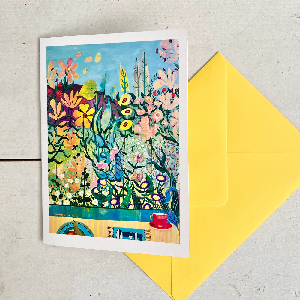 A colorful illustrated greeting card with a white border, placed on a light gray surface next to a bright yellow envelope. The card features a vibrant floral and garden scene with blue sky, green foliage, and multicolored flowers.