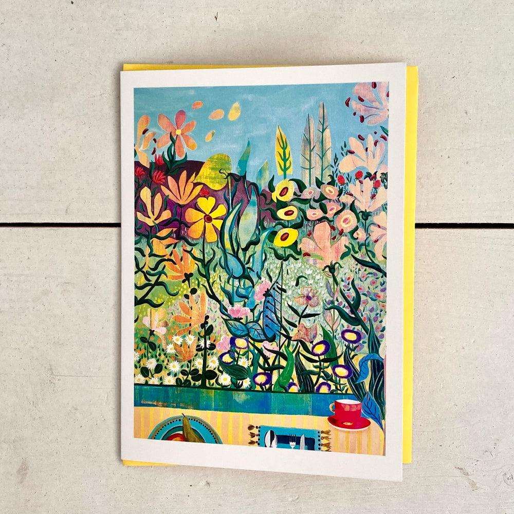 A colorful floral art print or card with a white border, leaning against a light gray textured background. The artwork features a dense, stylized garden scene with flowers, leaves, and birds in bright pastel and saturated tones, with a small red teacup illustration at the bottom.