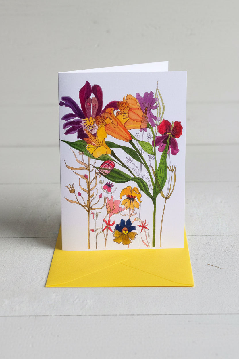 A vertical greeting card featuring a colorful botanical illustration of wildflowers, shown with a yellow envelope on a light gray wood-textured background.