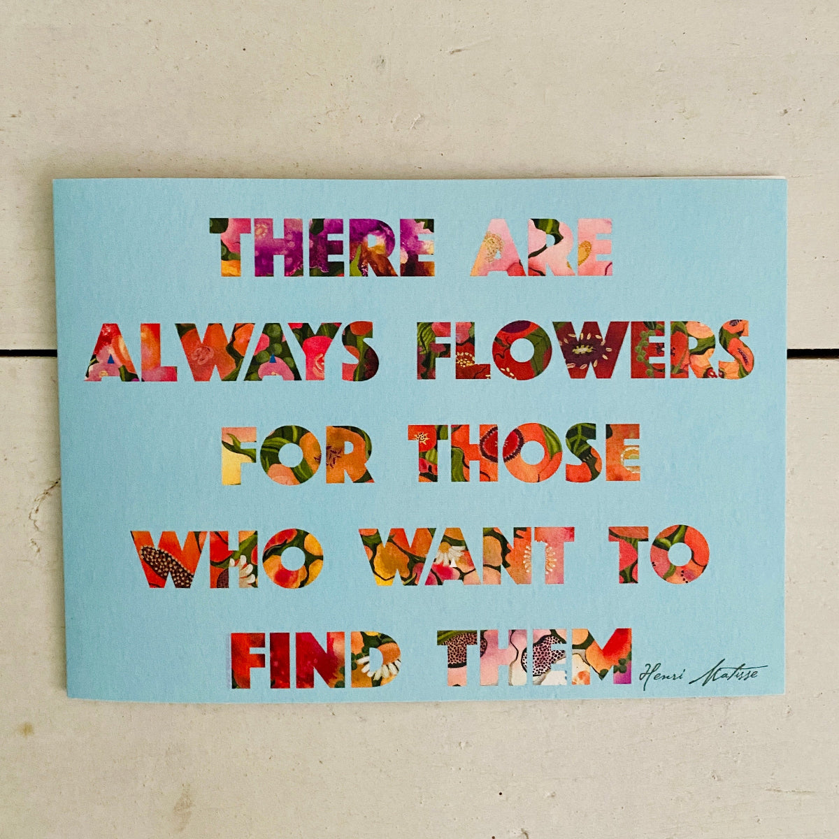 A square art print with a light blue background featuring the quote 'THERE ARE ALWAYS FLOWERS FOR THOSE WHO WANT TO FIND THEM' in colorful floral-patterned letters, displayed on a beige textured surface.