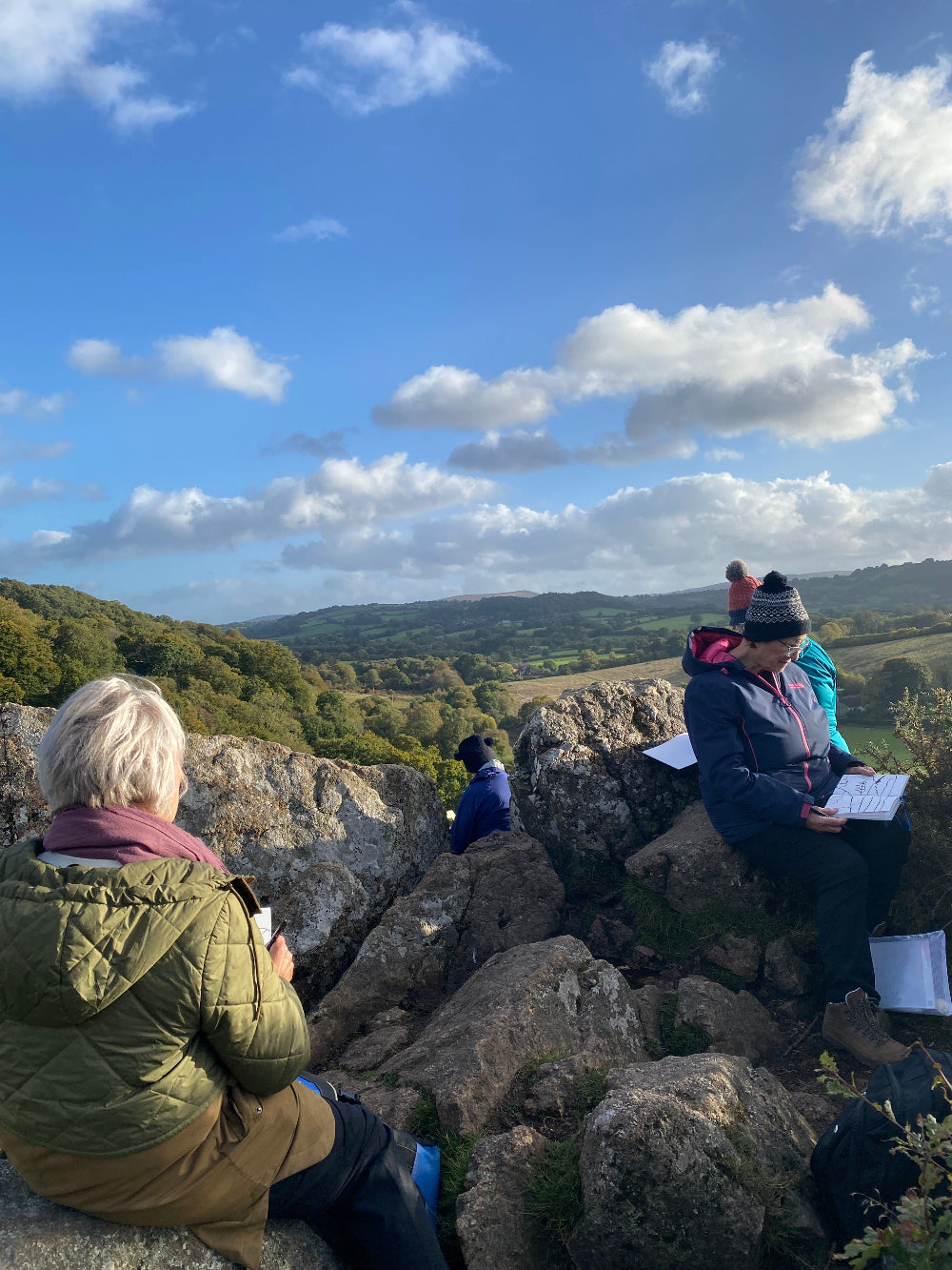 People sitting and sketching on a rocky cliff overlooking a scenic landscape with a clear blue sky.