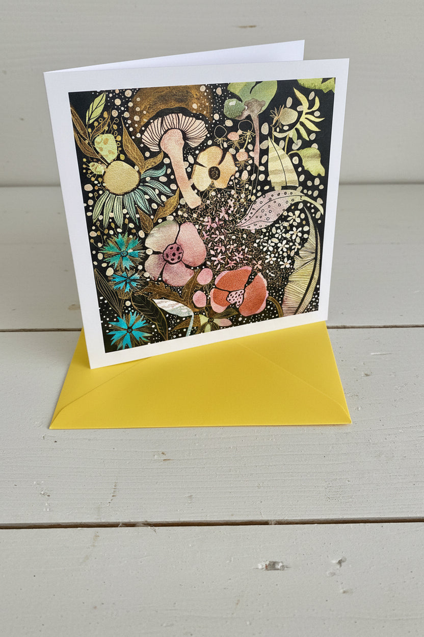 A square art print card with a white border featuring a dense, whimsical floral and mushroom illustration in black, gold, pink, blue, and green, shown with a bright yellow envelope on a white wood background.