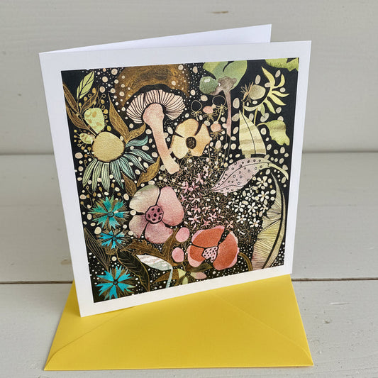 A square art print card with a white border featuring a dense, whimsical floral and mushroom illustration in black, gold, pink, blue, and green, shown with a bright yellow envelope on a white wood background.