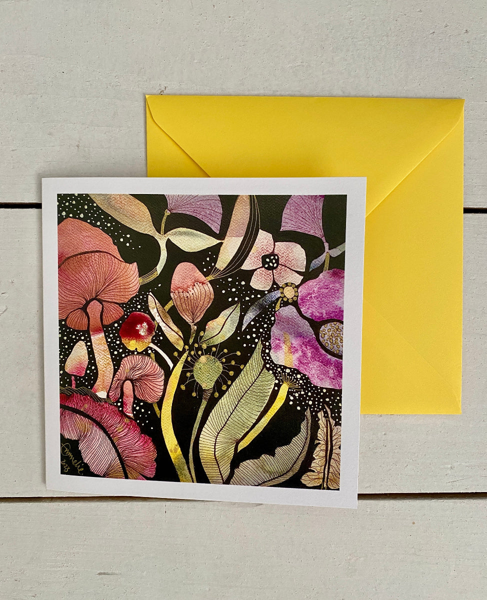 A square art print card featuring a colorful illustration of mushrooms and flowers on a black background, placed in front of a bright yellow envelope on a light wood surface.