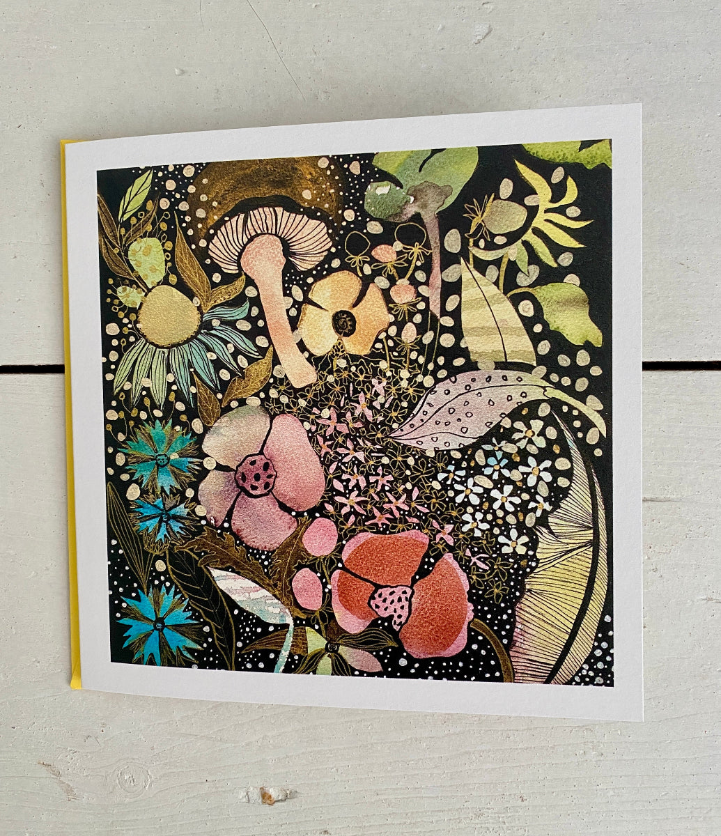 A square art print or card featuring a dense, whimsical floral and mushroom illustration on a black background, photographed from above on a light painted wood surface.