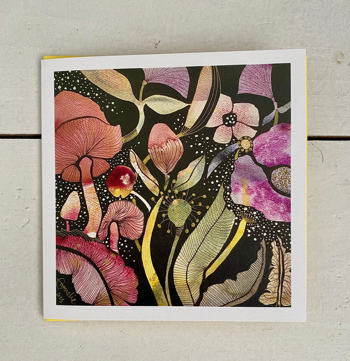 A square art print featuring a detailed botanical illustration of mushrooms and flowers in pink, purple, green, and gold tones on a black background, photographed flat on a light gray surface.