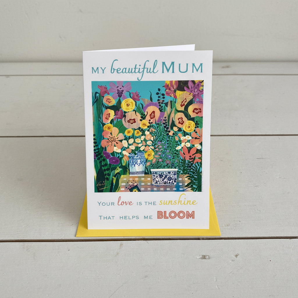 A portrait rectangular greeting card with a colourful floral illustration and the text 'MY beautiful MUM' and 'Your love is the sunshine THAT HELPS ME BLOOM', placed on top of a yellow envelope on a light grey wood plank background.