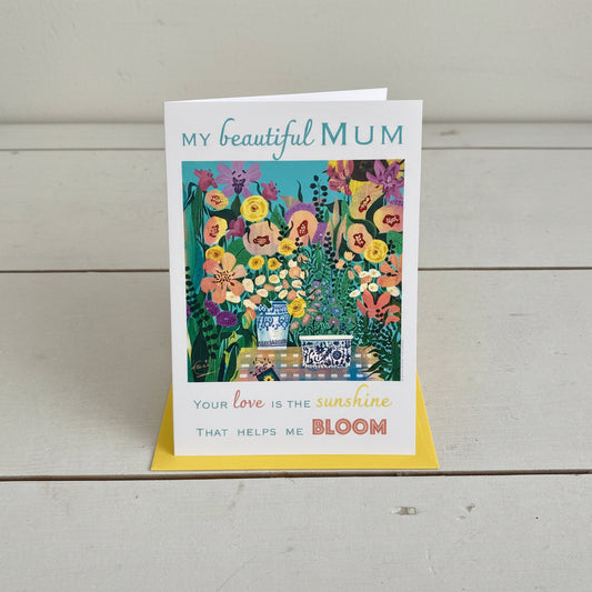 A portrait rectangular greeting card with a colourful floral illustration and the text 'MY beautiful MUM' and 'Your love is the sunshine THAT HELPS ME BLOOM', placed on top of a yellow envelope on a light grey wood plank background.