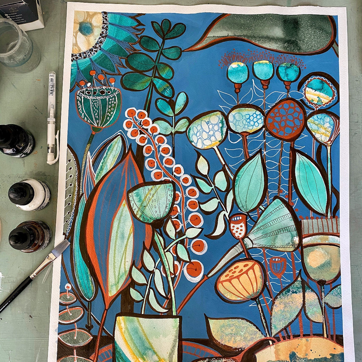 A colorful botanical art print featuring stylized leaves, flowers, and botanical motifs in teal, blue, orange, green, and cream, laid flat on a gray work surface with paint bottles, brushes, and a palette visible around it.