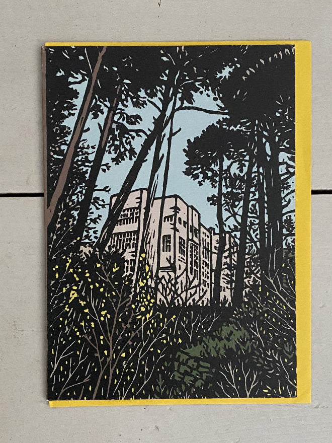 A vertical art print featuring a block-print style illustration of a building surrounded by dark trees and foliage, with a yellow border, displayed on a light gray wooden plank background.