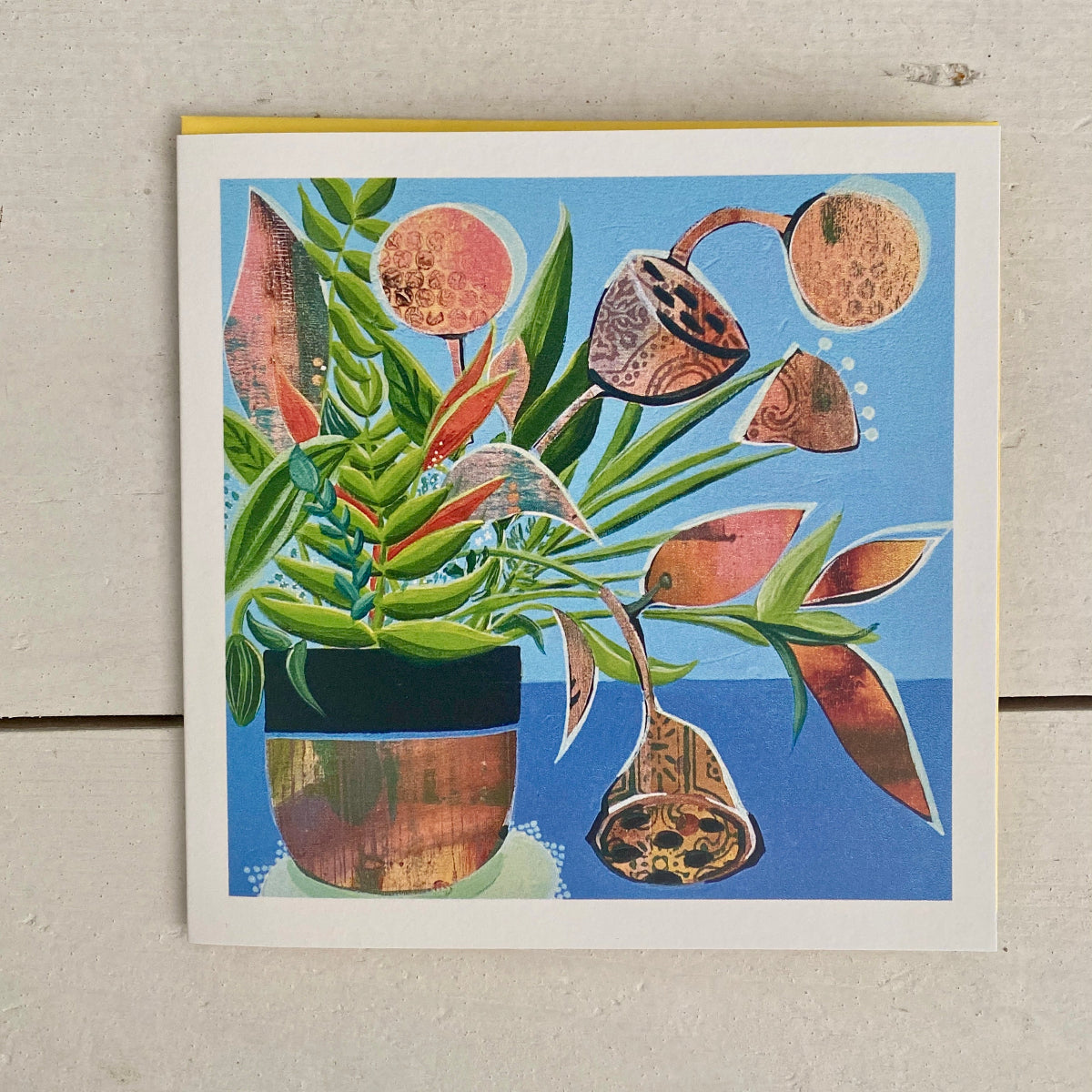 A square art print card, featuring a stylised botanical illustration of a potted plant with green leaves, copper-toned seed pods, and a blue background, photographed against a light textured wall.
