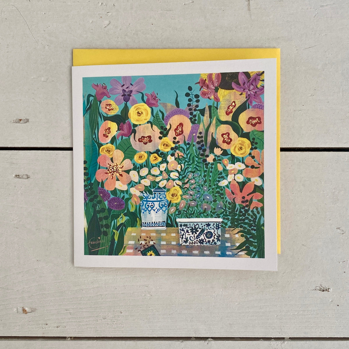 A square floral art print or greeting card with a colorful bouquet illustration, placed on top of a yellow envelope against a light weathered wood background.