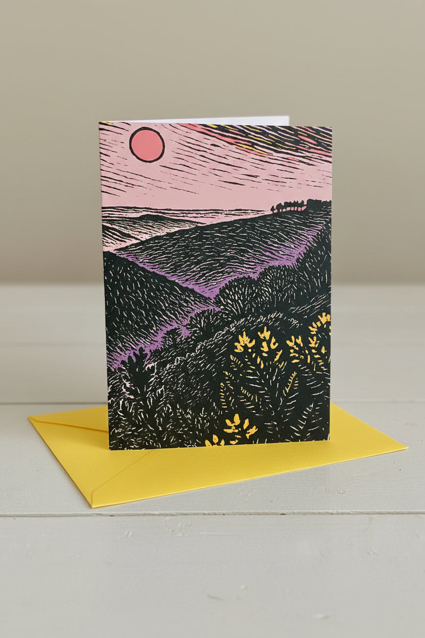 A rectangle  art print card featuring a stylized landscape illustration with a pink sky, red sun, black hills, and purple valley tones, placed over a bright yellow envelope on a light gray surface.