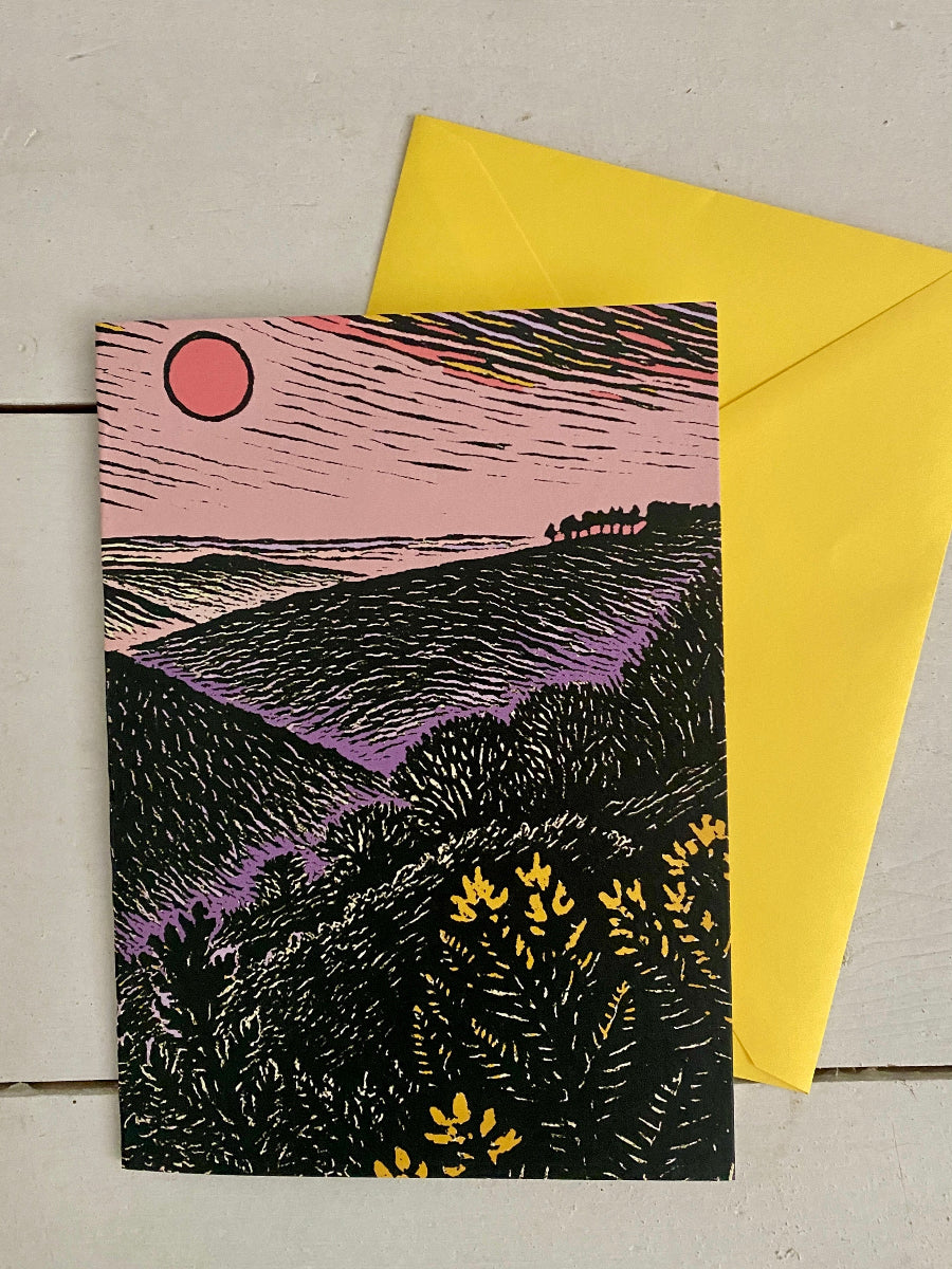 A square art print card featuring a stylized landscape illustration with a pink sky, red sun, black hills, and purple valley tones, placed over a bright yellow envelope on a light gray surface.