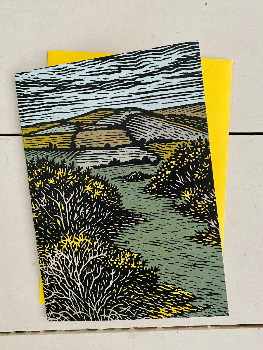 A landscape art print card featuring a block-print style illustration of a Welsh valley with gorse bushes, set against a bright yellow envelope on a light wood background.
