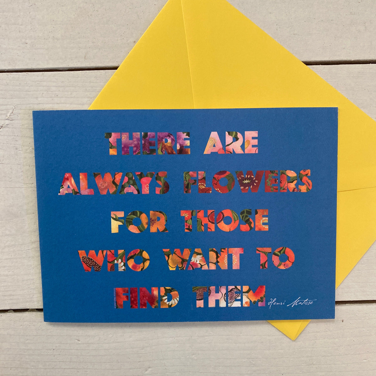 A blue greeting card with a floral-patterned quote reading 'THERE ARE ALWAYS FLOWERS FOR THOSE WHO WANT TO FIND THEM,' placed in front of a bright yellow envelope on a light wood surface.
