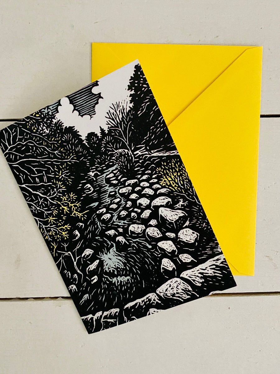 A black and white linocut-style art print of a stone path through trees, shown with a bright yellow envelope on a light wood background.