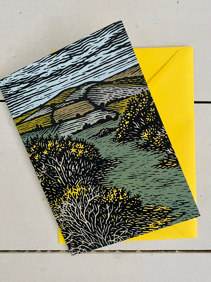 A landscape art print card featuring a stylized illustration of a Welsh countryside scene with yellow gorse and green hills, shown with a bright yellow envelope on a light wood background.