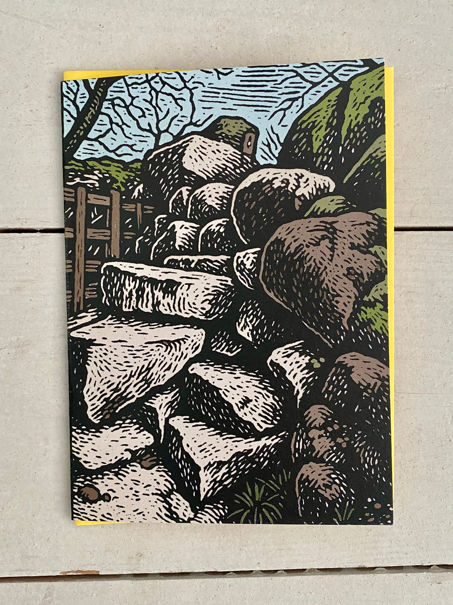 A rectangle art print or card featuring a block-print style illustration of stacked rocks, a wooden fence, and a blue sky, photographed flat on a light wood surface.