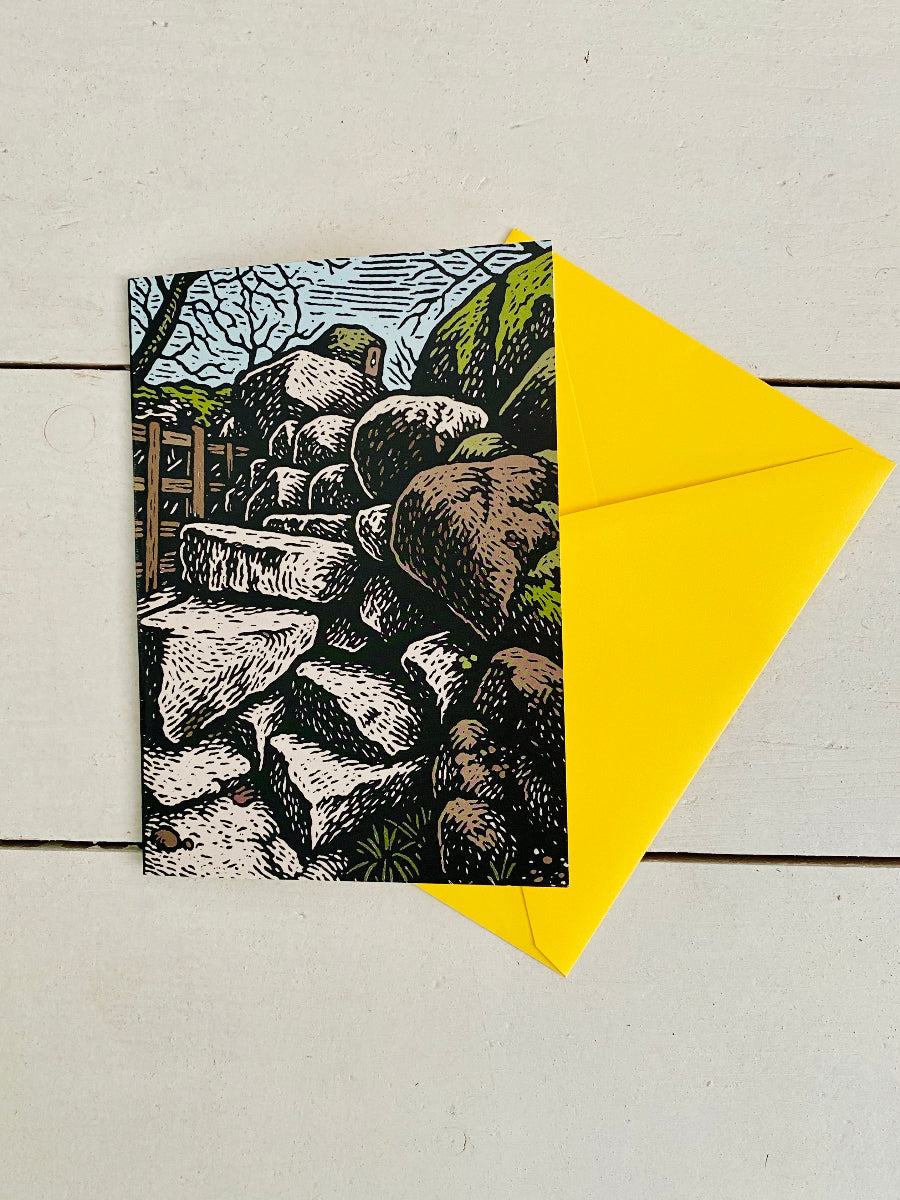A square art print or card featuring a black-and-white illustration of stacked rocks with green hills and a blue sky, placed on a white wood surface with a bright yellow envelope behind it.