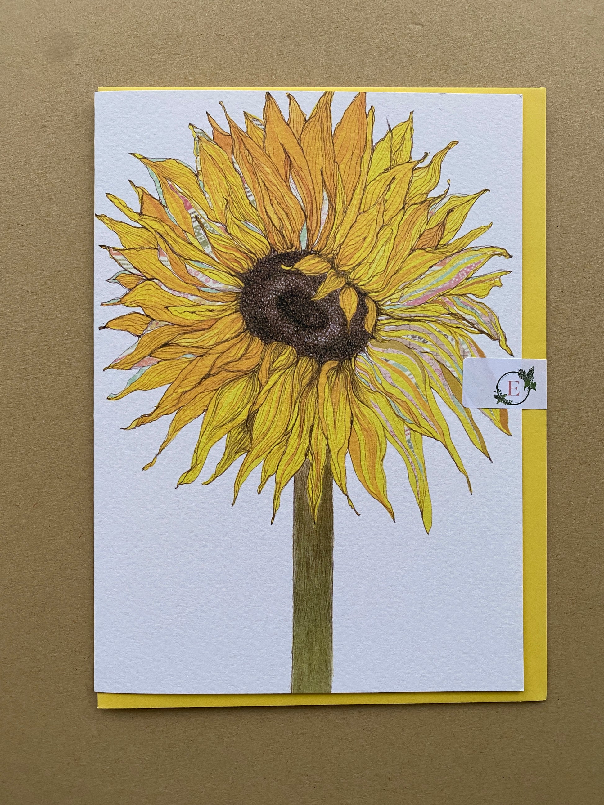 A square art print card featuring a detailed illustration of a yellow sunflower, set against a white background with a yellow envelope displayed on a textured brown surface. With a paper clasp sticker.