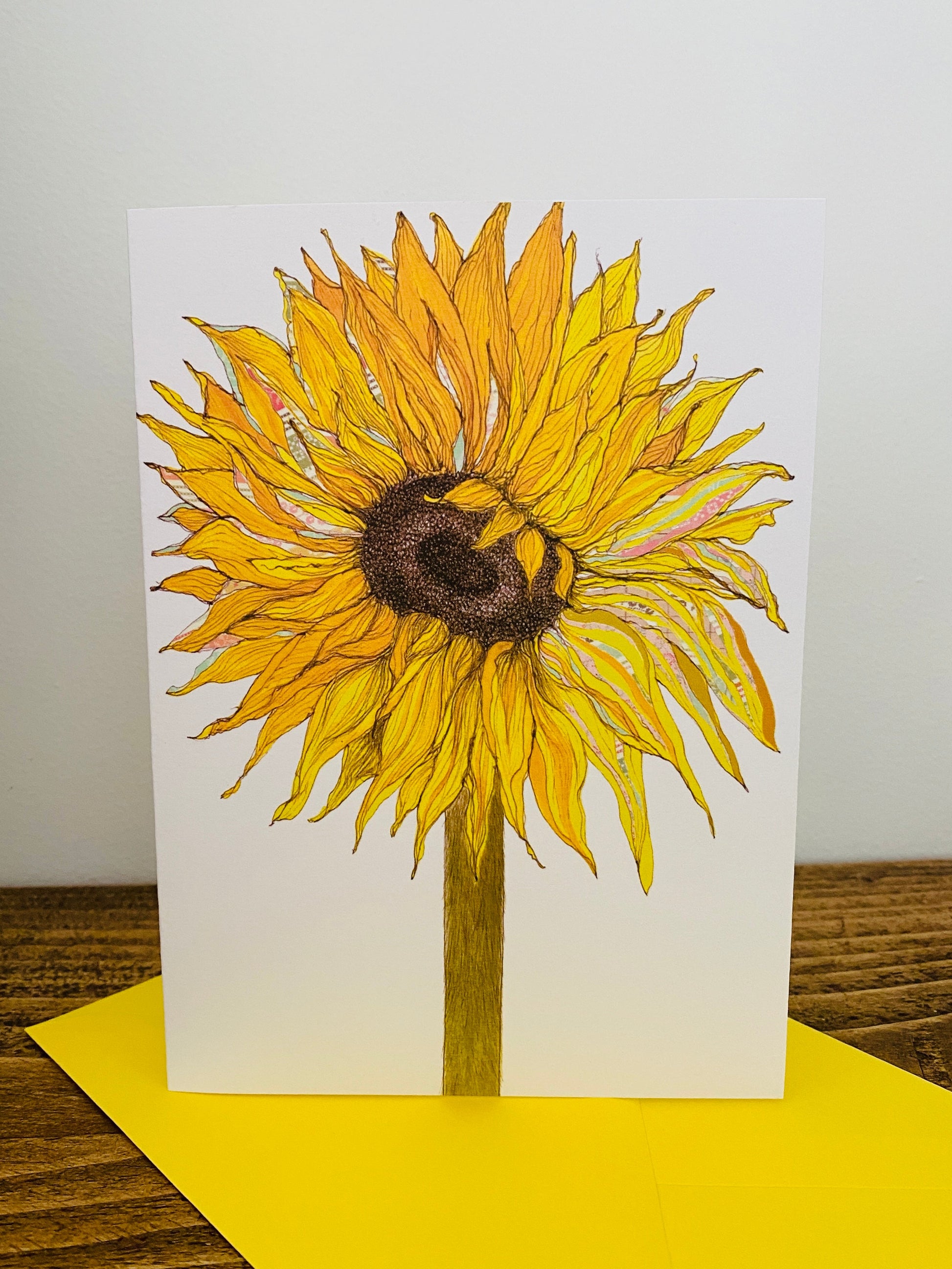 A vertical white greeting card featuring a hand-drawn yellow sunflower with a brown center and green stem, standing upright on a yellow envelope over a wooden surface against a light gray wall.