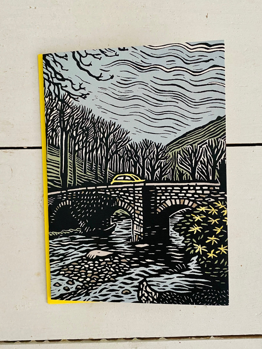 A square art print featuring a black and white linocut-style illustration of a stone bridge over a river, with bare trees, hills, and a yellow car on the bridge. The print is photographed flat against a light beige textured background.