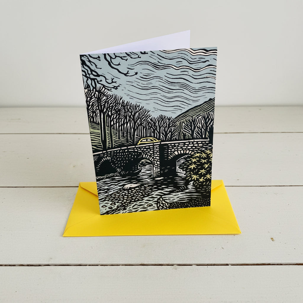 A rectangle art print card featuring a black and white linocut-style illustration of a stone bridge over water, with a bright yellow envelope partially visible behind it on a light wood surface.