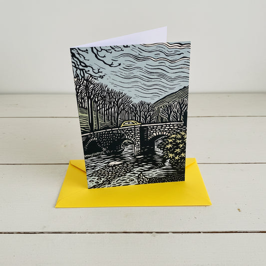 A rectangle art print card featuring a black and white linocut-style illustration of a stone bridge over water, with a bright yellow envelope partially visible behind it on a light wood surface.