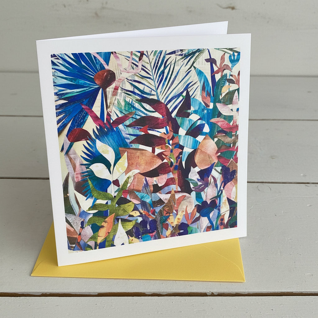 A square art card, featuring a colourful abstract botanical pattern with layered leaves and floral shapes, displayed on a light grey wood surface with a yellow card or paper peeking out from beneath.