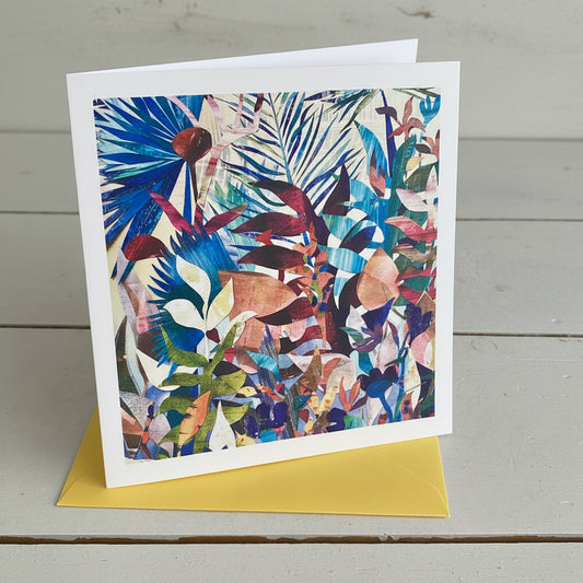 A square art card, featuring a colourful abstract botanical pattern with layered leaves and floral shapes, displayed on a light grey wood surface with a yellow card or paper peeking out from beneath.