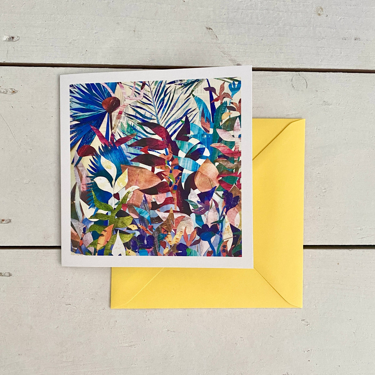A square art card print with a colourful abstract botanical pattern, displayed on top of a bright yellow envelope on a light grey wood plank background.