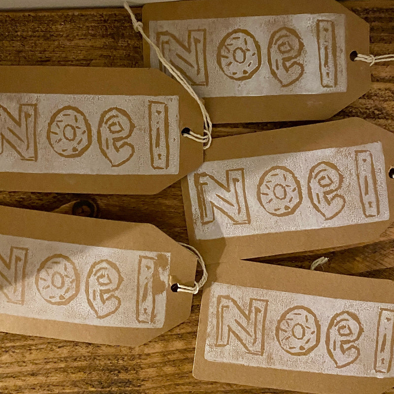 Brown paper tags with 'Noel' printed on them, hanging on a wooden surface.