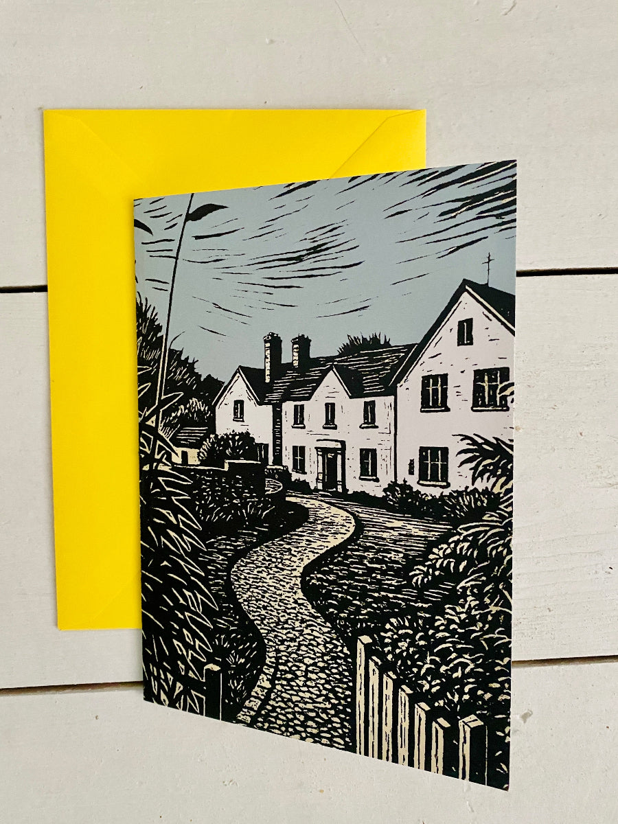 A square art print or card featuring a black-and-white linocut-style illustration of a cottage and winding path, shown with a bright yellow envelope or backing card on a white wood surface.