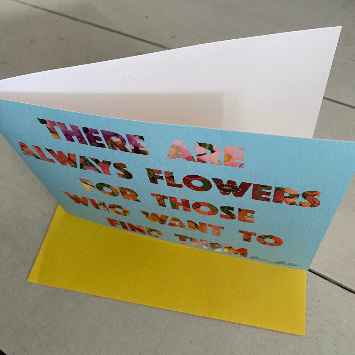 A folded greeting card with a light blue front, white interior, and yellow backing, printed with the phrase 'THERE ARE ALWAYS FLOWERS FOR THOSE WHO WANT TO FIND THEM' in multicolored lettering, photographed on a light floor.