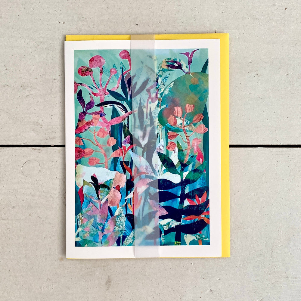 An abstract botanical art print with vibrant pink, teal, and green brushstrokes, presented on a white cardstock with a yellow envelope, set against a light gray wood background.