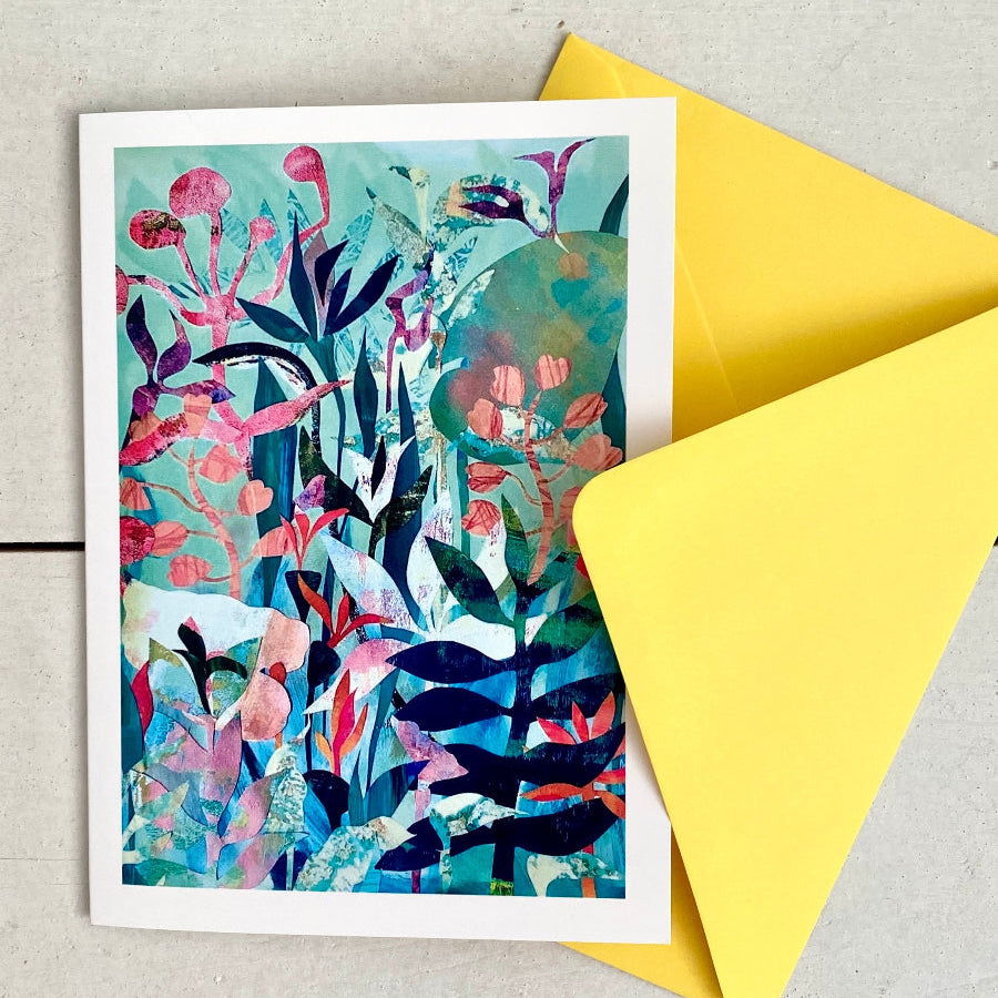 A botanical art print card with a white border, featuring a colorful abstract floral and leaf design in teal, pink, and navy, shown with a bright yellow envelope on a light gray wood background.