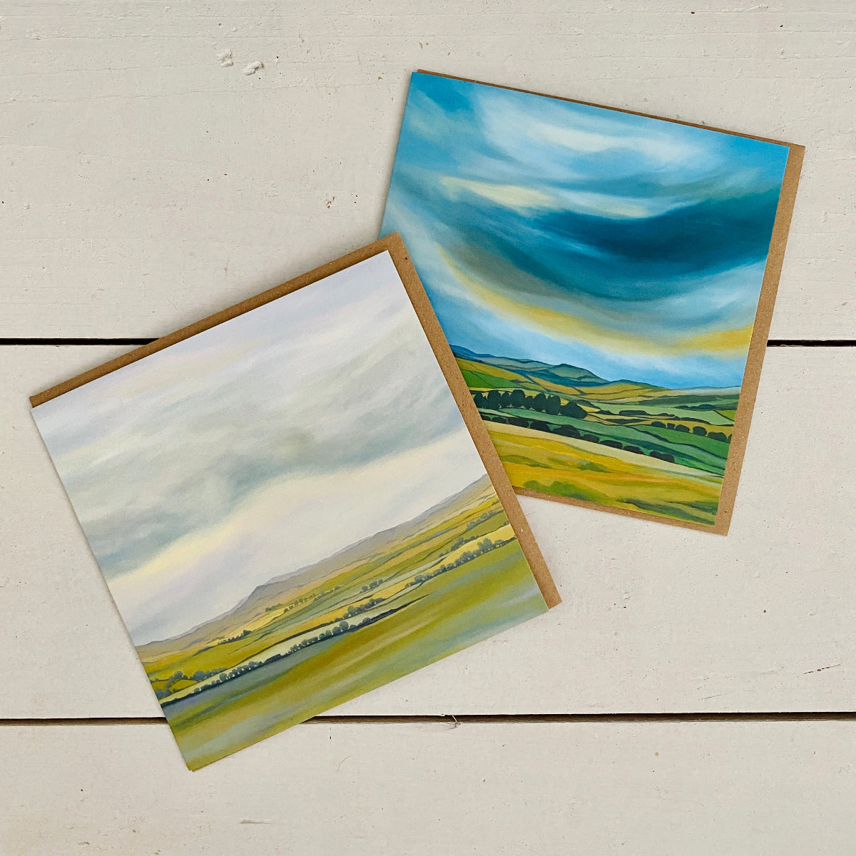 Two square landscape paintings with brown paper borders, displayed diagonally on a white wooden surface. One painting depicts rolling green and yellow fields under a cloudy sky, while the other shows dramatic blue and yellow clouds over green hills.