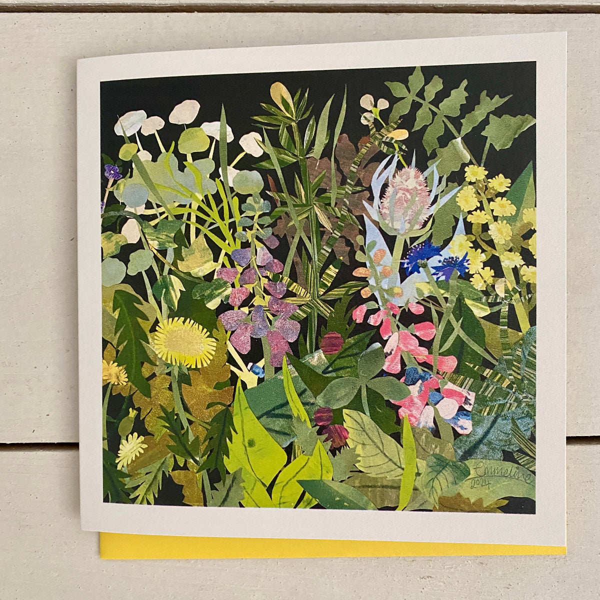 A square art print card featuring a dense, colourful wildflower and mushroom illustration, displayed flat on a light beige wood surface with a yellow card peeking out from underneath.