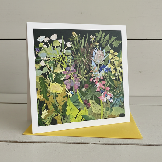 A square art print or greeting card with a white border, featuring a colorful botanical illustration of wildflowers and green leaves on a dark background, displayed upright on a yellow envelope against a light gray wood plank surface.