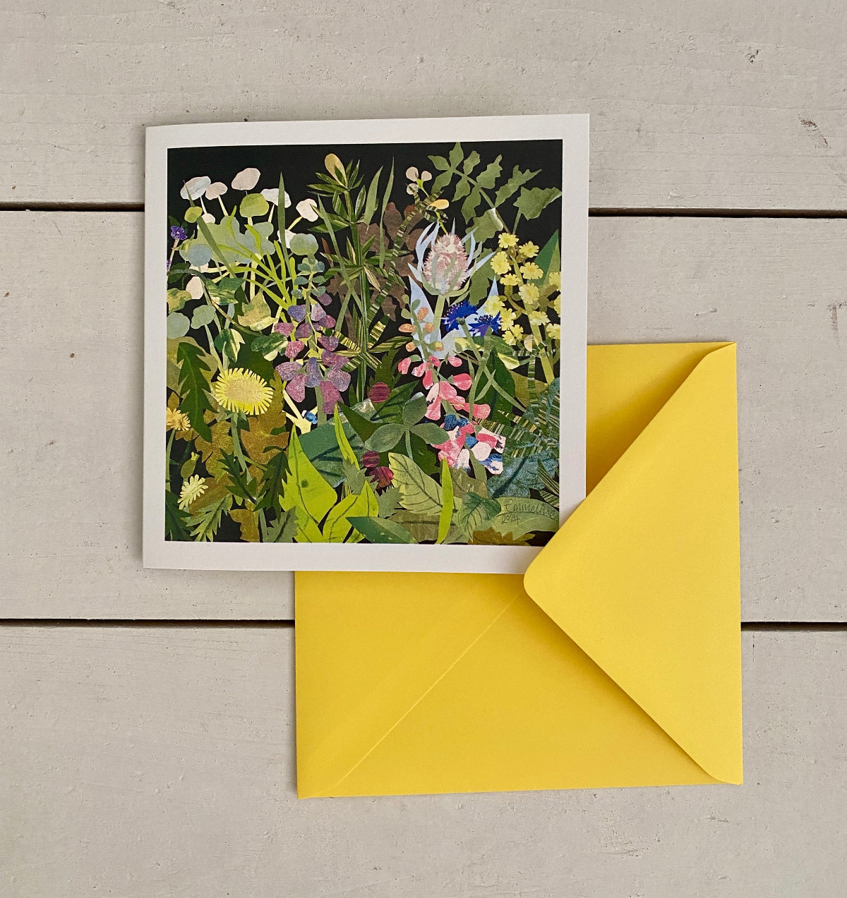 A square floral art print card, with a white border, placed on a light wood surface next to a bright yellow envelope. The artwork depicts a dense arrangement of wildflowers with green foliage and a dark background.