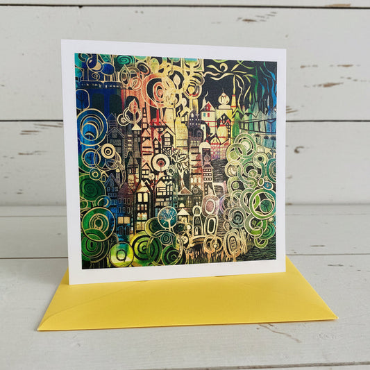 A square art print card with a white border showing an abstract cityscape illustration in black, green, yellow, orange, and blue, placed in front of a bright yellow envelope on a light wood surface.